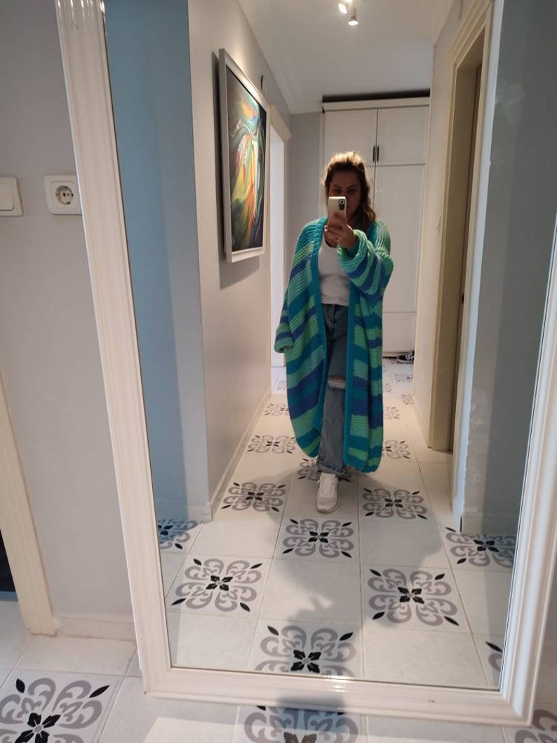 May include: A long, open-front cardigan in shades of blue and green, with horizontal stripes. The cardigan is being worn in a hallway with a decorative tile floor and a large mirror.