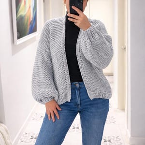 Puede incluir: Un c&aacute;rdigan de punto grueso en color gris claro con un patr&oacute;n de textura, dise&ntilde;o frontal abierto y mangas largas voluminosas con pu&ntilde;os acanalados. La prenda se lleva sobre una camiseta negra de cuello alto y pantalones vaqueros azules.
