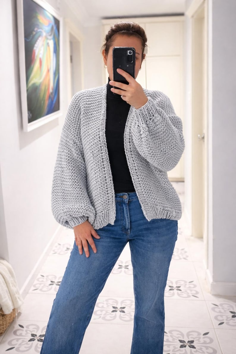 Puede incluir: Un c&aacute;rdigan de punto grueso en color gris claro con un patr&oacute;n de textura, dise&ntilde;o frontal abierto y mangas largas voluminosas con pu&ntilde;os acanalados. La prenda se lleva sobre una camiseta negra de cuello alto y pantalones vaqueros azules.