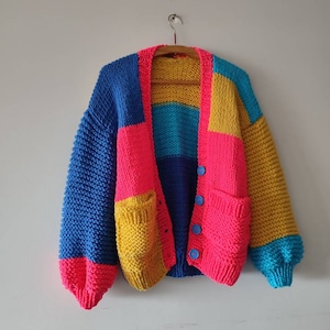 May include: A colourful, chunky knit cardigan with a patchwork design. The sweater features blocks of blue, pink, yellow, and teal. It has two front pockets and is fastened with blue buttons. The cardigan is displayed on a wooden hanger.