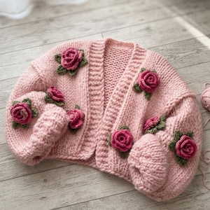 Op de afbeelding: Een grof gebreid vest in een zachte pastelroze kleur, met een open voorzijde en lange mouwen. Het kledingstuk is versierd met diverse driedimensionale gehaakte rozenapplicaties in donkerroze tinten met groene bladeren.
