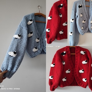 K&ouml;nnte beinhalten: Hellblau und rote Strickjacke mit wei&szlig;en und schwarzen Schafapplikationen. Die Strickjacke hat lange &Auml;rmel und einen verk&uuml;rzten Schnitt. Die Schafe sind gleichm&auml;&szlig;ig &uuml;ber dem Kleidungsst&uuml;ck verteilt.