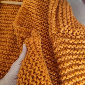 Puede incluir: Un c&aacute;rdigan de punto grueso en un vibrante color naranja mostaza. La prenda presenta un patr&oacute;n de punto bobo texturizado y denso en toda su superficie, con un cuello ancho y mangas largas, destacando la construcci&oacute;n de lana tejida a mano.