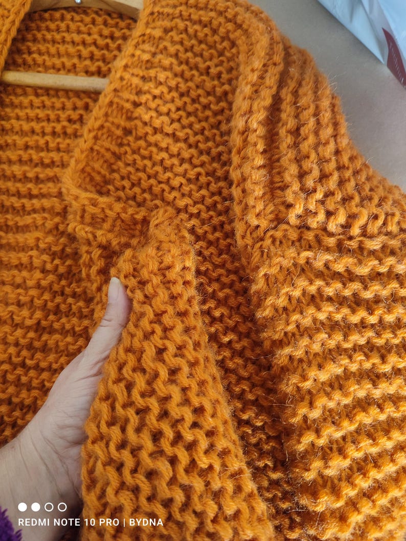 Puede incluir: Un c&aacute;rdigan de punto grueso en un vibrante color naranja mostaza. La prenda presenta un patr&oacute;n de punto bobo texturizado y denso en toda su superficie, con un cuello ancho y mangas largas, destacando la construcci&oacute;n de lana tejida a mano.