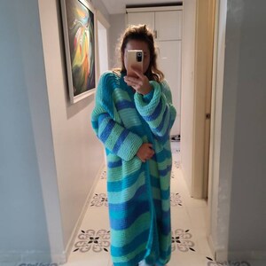 May include: A long, open-front cardigan in shades of blue and green. The knitted garment features horizontal stripes and long sleeves. The cardigan appears to be handmade and is suitable for layering. The style is casual and comfortable.