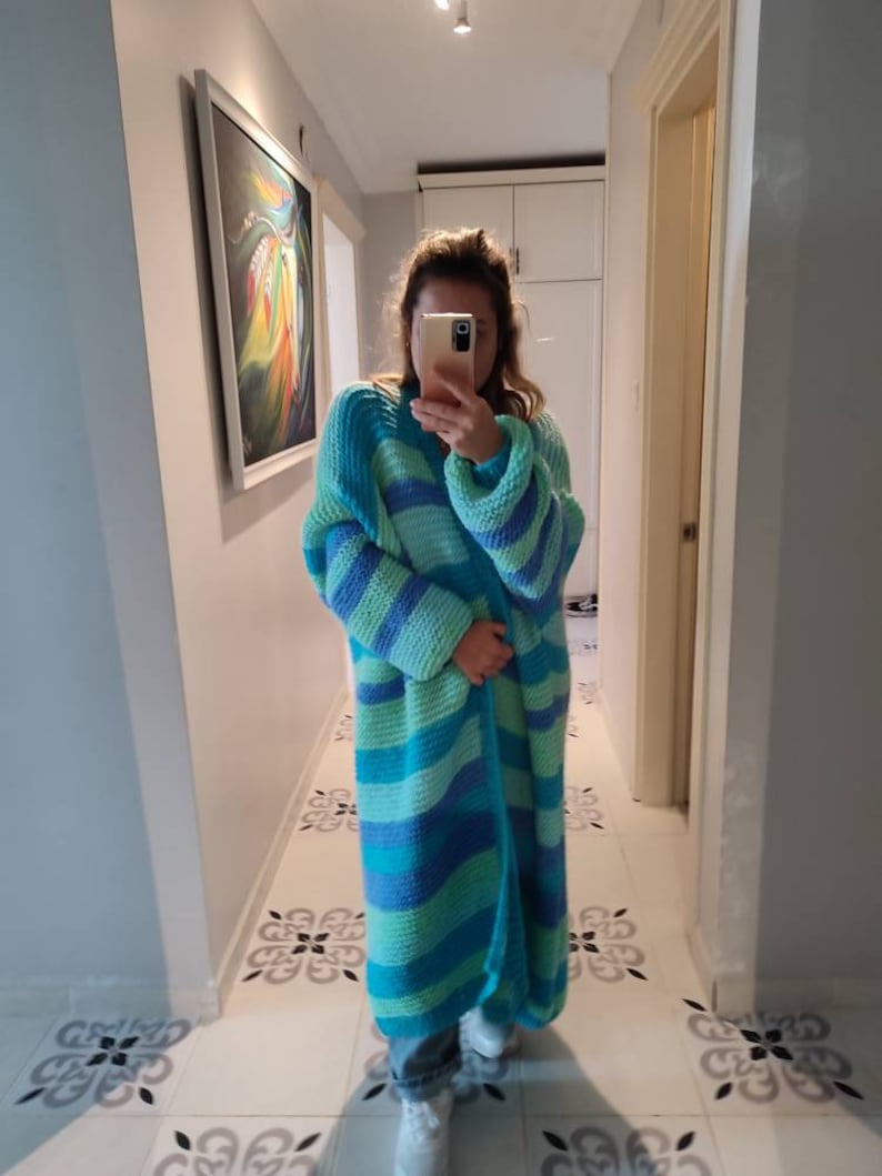 May include: A long, open-front cardigan in shades of blue and green. The knitted garment features horizontal stripes and long sleeves. The cardigan appears to be handmade and is suitable for layering. The style is casual and comfortable.