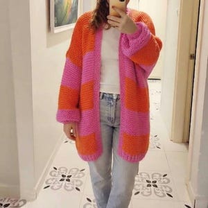 May include: A chunky knit cardigan featuring wide horizontal stripes in vibrant orange and bright pink. The open-front garment has long sleeves with dropped shoulders and a relaxed fit, worn over a plain white t-shirt and light blue denim jeans.