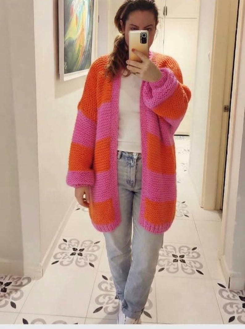May include: A chunky knit cardigan featuring wide horizontal stripes in vibrant orange and bright pink. The open-front garment has long sleeves with dropped shoulders and a relaxed fit, worn over a plain white t-shirt and light blue denim jeans.