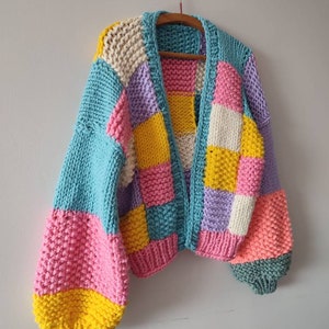 Könnte beinhalten: Ein grob gestrickter Cardigan im Patchwork-Design. Der Pullover hat eine Mischung aus Pastellfarben, darunter Blau, Rosa, Gelb, Lila und Weiß. Die Ärmel sind einfarbig blau und rosa, und der Cardigan hängt an einem Holzbügel.