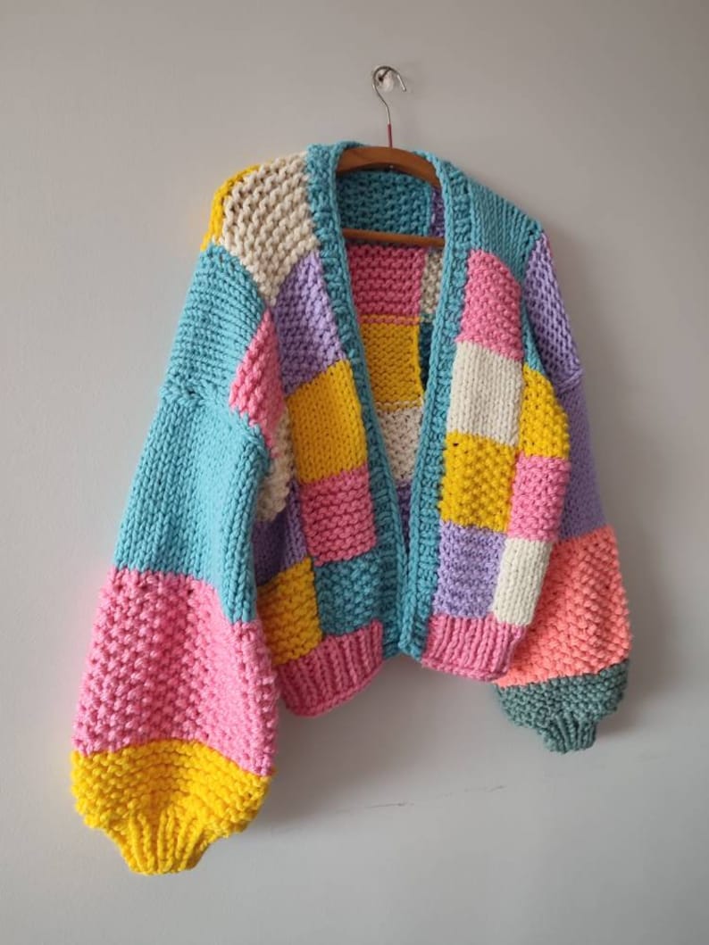 May include: A chunky knit cardigan in a patchwork design. The sweater features a mix of pastel colors, including blue, pink, yellow, purple, and white. The sleeves are a solid blue and pink, and the cardigan is hanging on a wooden hanger.