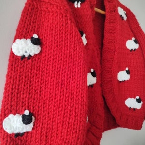 K&ouml;nnte beinhalten: Ein leuchtend roter, grob gestrickter Cardigan mit Applikationen von wei&szlig;en und schwarzen Schafen. Der gem&uuml;tliche Pullover hat eine Knopfleiste vorne und lange &Auml;rmel. Die Schafe sind gleichm&auml;&szlig;ig &uuml;ber Vorder- und &Auml;rmel verteilt.