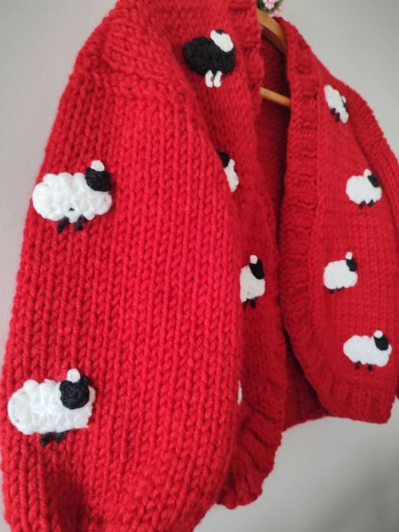 K&ouml;nnte beinhalten: Ein leuchtend roter, grob gestrickter Cardigan mit Applikationen von wei&szlig;en und schwarzen Schafen. Der gem&uuml;tliche Pullover hat eine Knopfleiste vorne und lange &Auml;rmel. Die Schafe sind gleichm&auml;&szlig;ig &uuml;ber Vorder- und &Auml;rmel verteilt.