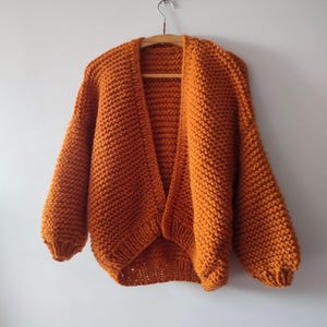 Puede incluir: Un c&aacute;rdigan de punto grueso en un vibrante color naranja quemado. Esta prenda abierta en la parte delantera presenta mangas largas y voluminosas con pu&ntilde;os acanalados y un patr&oacute;n de punto bobo texturizado en toda su superficie.