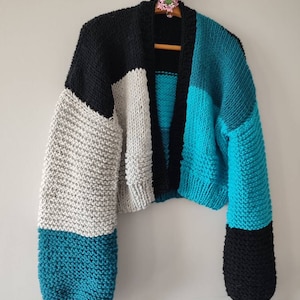 May include: A chunky knit cardigan in a color block design. The sweater features black, gray, and teal panels. The sleeves are long and the body is cropped. The cardigan is displayed on a wooden hanger.