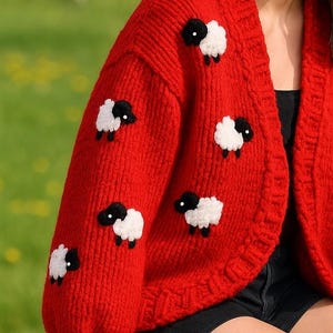 K&ouml;nnte beinhalten: Ein leuchtend roter, grob gestrickter Cardigan mit wei&szlig;en Schafen mit schwarzen Gesichtern. Der Cardigan mit offenem Schnitt hat eine lockere Passform. Die Schafe sind gleichm&auml;&szlig;ig &uuml;ber die &Auml;rmel und die Vorderseite verteilt.