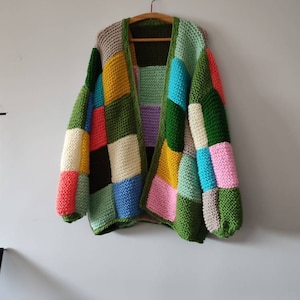 May include: A handmade, patchwork cardigan in a variety of colors, including green, yellow, blue, pink, and black. The sweater is hanging on a wooden hanger against a white wall. The knitted garment has a chunky texture.