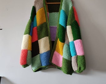 Puede incluir: Un cárdigan patchwork hecho a mano en una variedad de colores, incluyendo verde, amarillo, azul, rosa y negro. El suéter está colgado en una percha de madera contra una pared blanca. La prenda de punto tiene una textura gruesa.