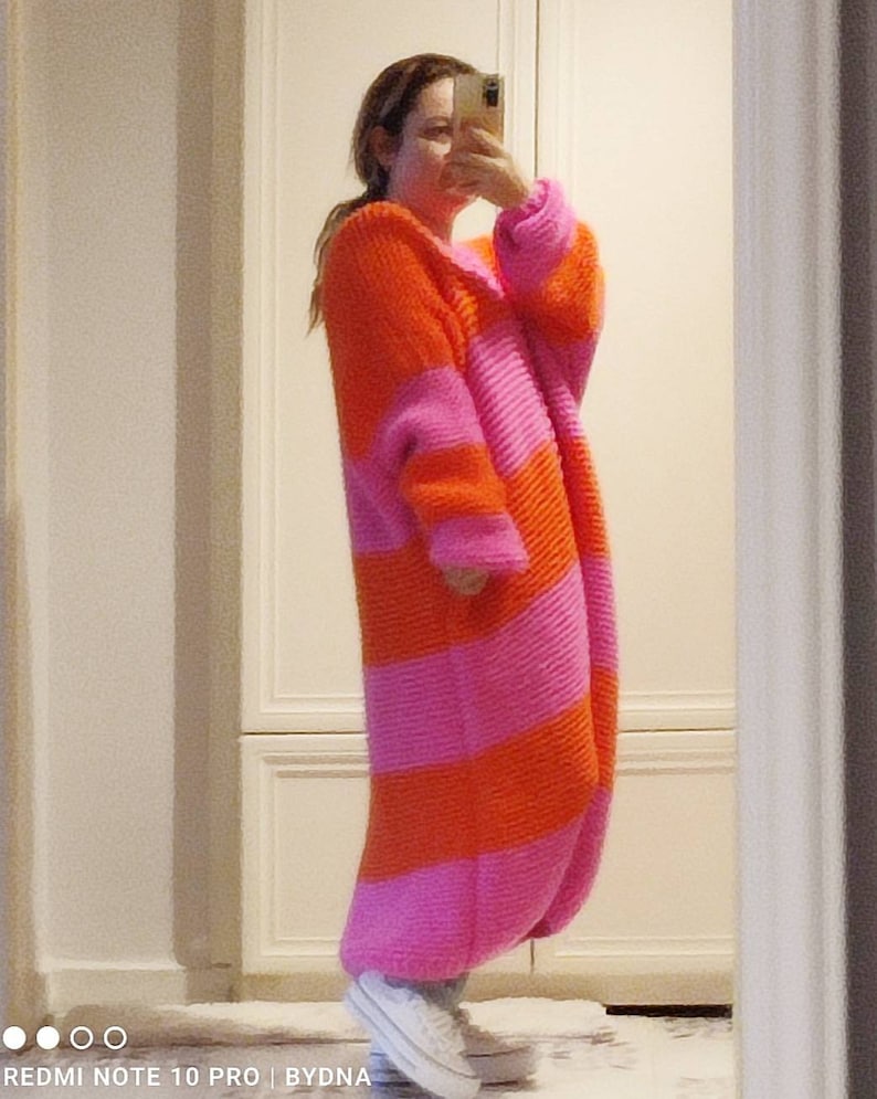May include: A long-sleeved, ankle-length knit cardigan featuring wide, horizontal stripes in alternating bright orange and vibrant pink. The garment has a thick, textured yarn construction with a relaxed fit, long sleeves, and a simple open front design.