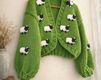 May include: A green, cropped cardigan sweater with a sheep design. The sweater is made of a chunky knit and features white and black sheep appliqués. The sweater is hanging on a wooden hanger against a neutral background.