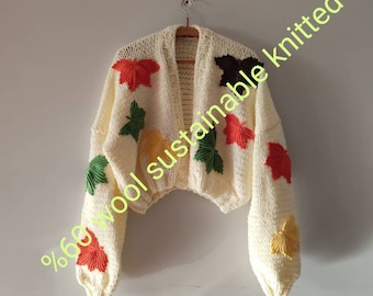 May include: Cream-coloured, knitted cardigan with colourful leaf appliqués. The open-front sweater features red, green, yellow, and brown leaves. The text on the image reads: "60% wool sustainable knitted".