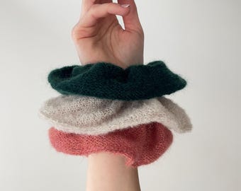 May include: Three knitted hair scrunchies in a stack. The bottom scrunchie is a light pink color, the middle scrunchie is a light beige color, and the top scrunchie is a dark green color.