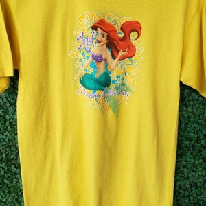 May include: Yellow t-shirt with a graphic of Ariel, the Little Mermaid, sitting on a bed of flowers. The text "Ariel Princess of the Sea" is printed below the image.