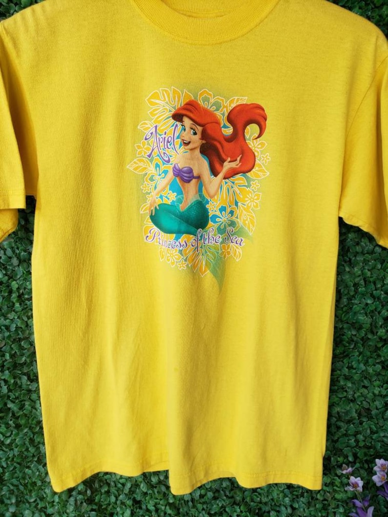 May include: Yellow t-shirt with a graphic of Ariel, the Little Mermaid, sitting on a bed of flowers. The text "Ariel Princess of the Sea" is printed below the image.