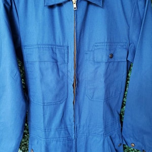 May include: A blue denim work jumpsuit with a zipper closure and two chest pockets. The jumpsuit has a brand label that reads "Wrangler" on the front.