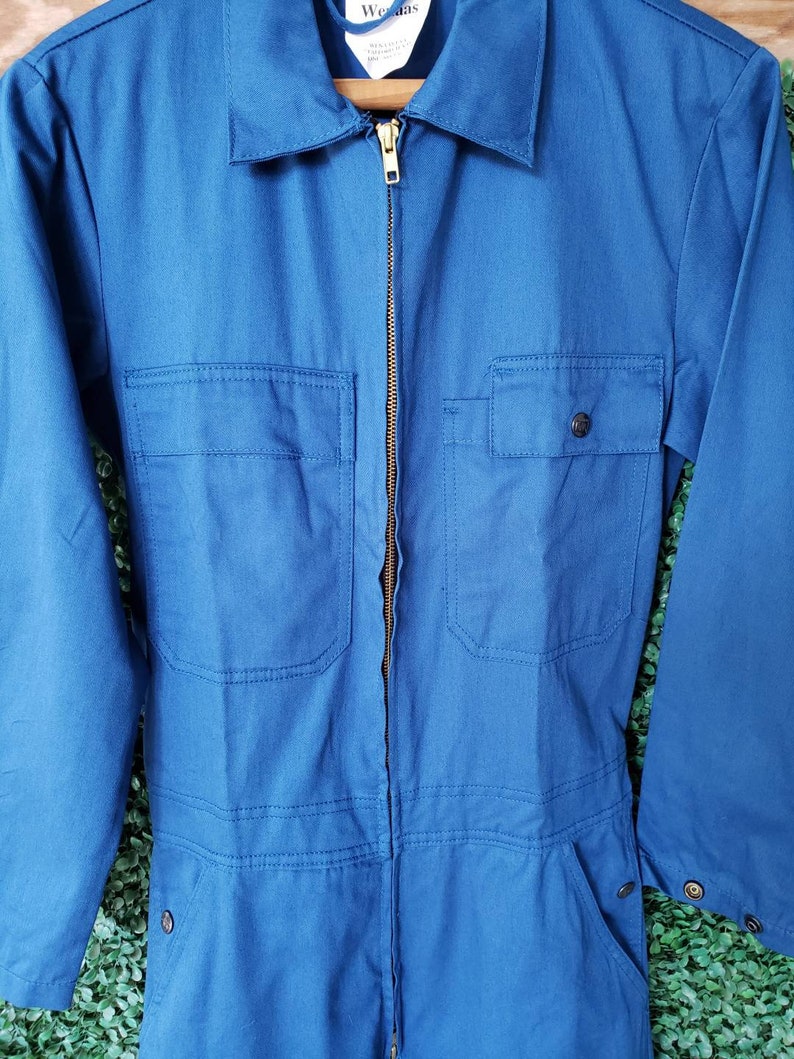May include: A blue denim work jumpsuit with a zipper closure and two chest pockets. The jumpsuit has a brand label that reads "Wrangler" on the front.