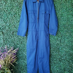 May include: A blue denim jumpsuit with a zipper closure and multiple pockets. The jumpsuit has long sleeves and a collared neckline.