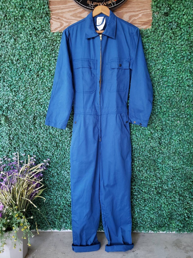 May include: A blue denim jumpsuit with a zipper closure and multiple pockets. The jumpsuit has long sleeves and a collared neckline.