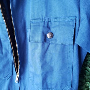 May include: A blue workwear jumpsuit with a zipper closure and a pocket on the chest. The pocket has a metal button with a logo.