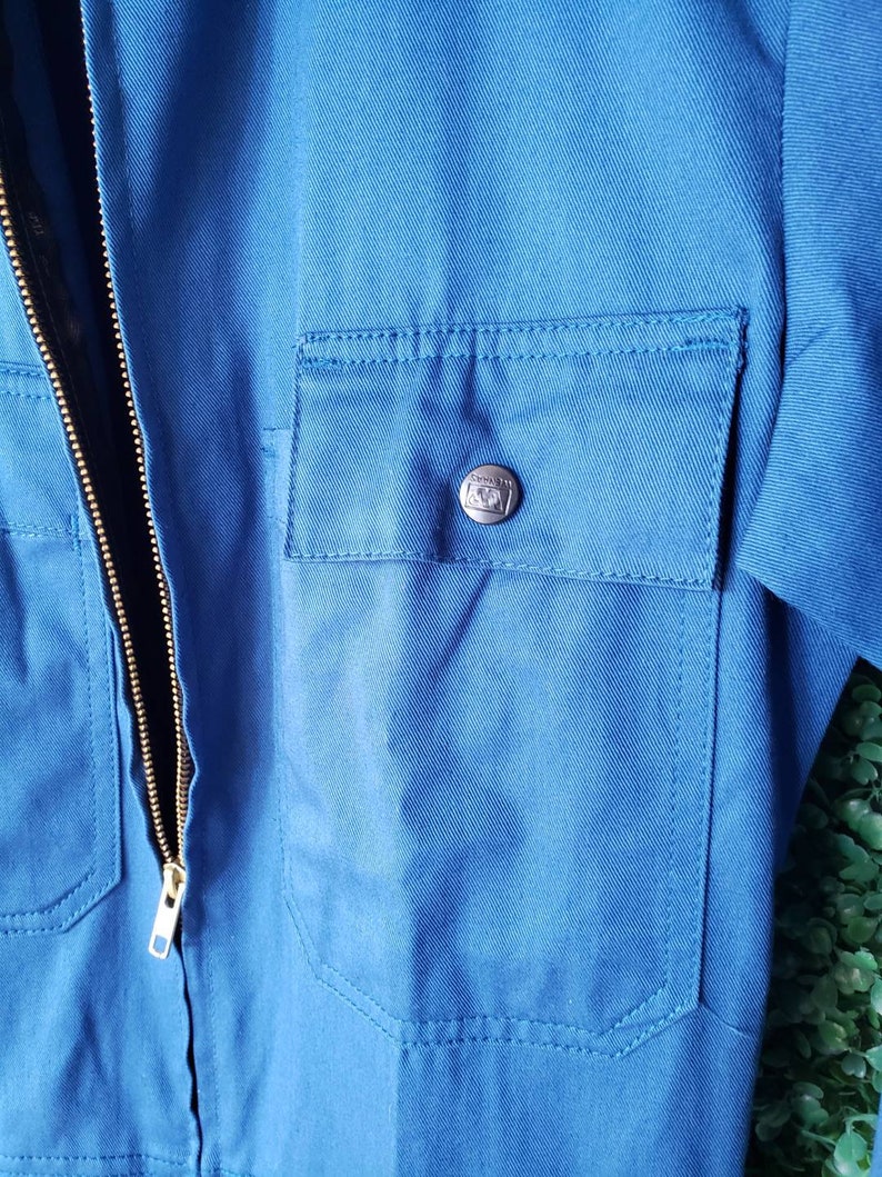 May include: A blue workwear jumpsuit with a zipper closure and a pocket on the chest. The pocket has a metal button with a logo.