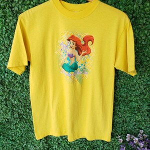 May include: Yellow t-shirt featuring a graphic of Ariel, the Little Mermaid, with the text "Ariel, Princess of the Sea".