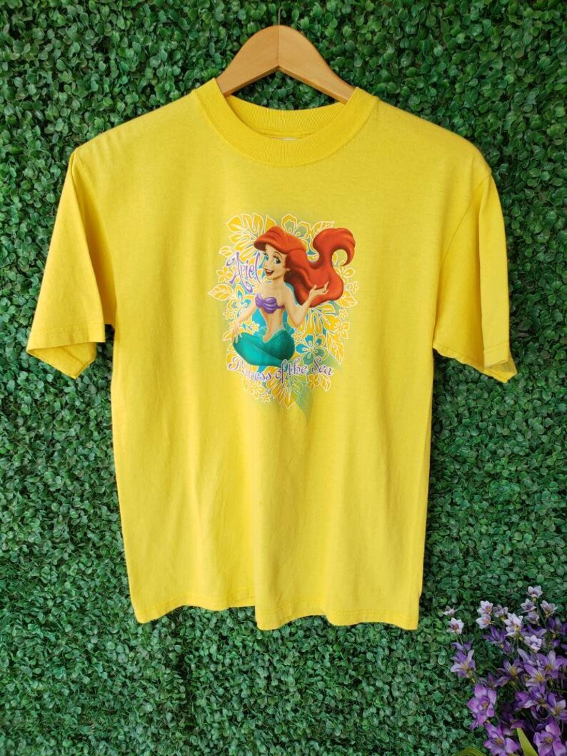May include: Yellow t-shirt featuring a graphic of Ariel, the Little Mermaid, with the text "Ariel, Princess of the Sea".