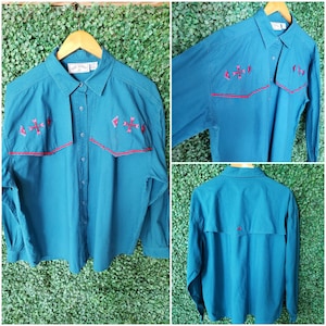 May include: Teal blue long-sleeve button-up shirt with red and white embroidered geometric designs on the shoulders and back. The shirt has a pointed collar and a single button closure at the top.