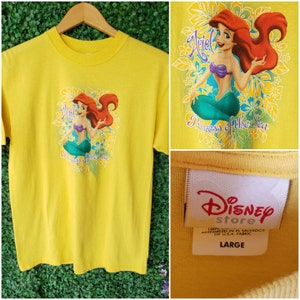 May include: Yellow t-shirt featuring a cartoon image of Ariel, the Little Mermaid, with the text "Princess of the Sea". The shirt has a Disney Store tag with the text "100% Cotton, Assembled in El Salvador of U.S.A. Fabric, Large".