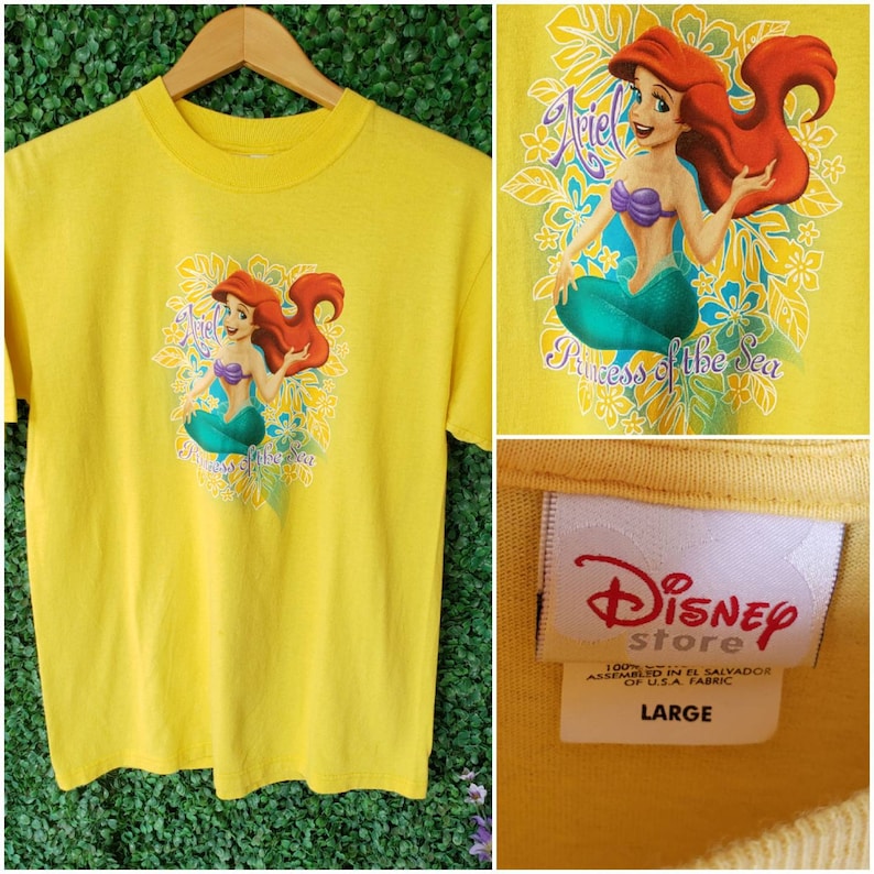 May include: Yellow t-shirt featuring a cartoon image of Ariel, the Little Mermaid, with the text "Princess of the Sea". The shirt has a Disney Store tag with the text "100% Cotton, Assembled in El Salvador of U.S.A. Fabric, Large".