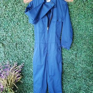 May include: A blue denim jumpsuit with a zipper closure and two pockets on the chest. The jumpsuit has long sleeves and is cuffed at the ankles.