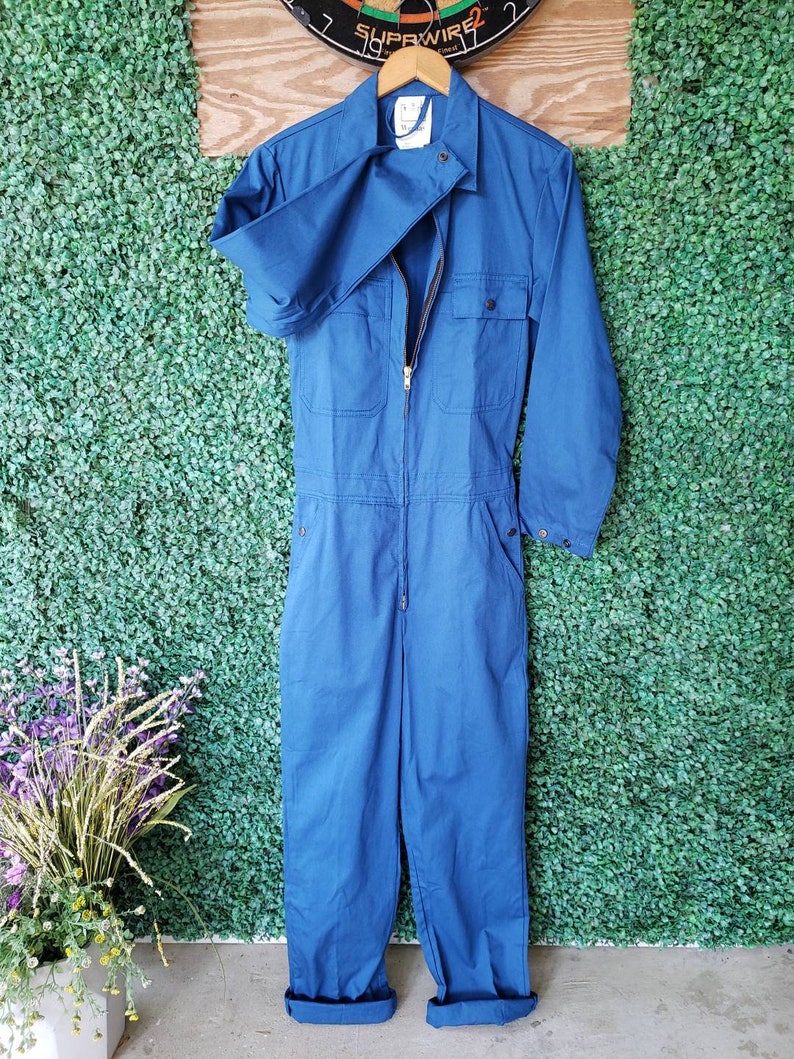 May include: A blue denim jumpsuit with a zipper closure and two pockets on the chest. The jumpsuit has long sleeves and is cuffed at the ankles.