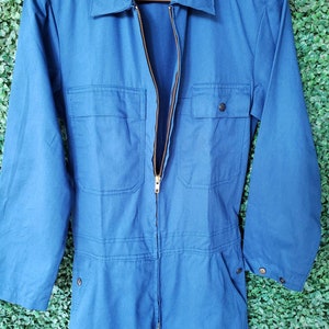 May include: A blue denim jumpsuit with a zipper closure and two pockets on the front. The jumpsuit has long sleeves and a collared neckline.