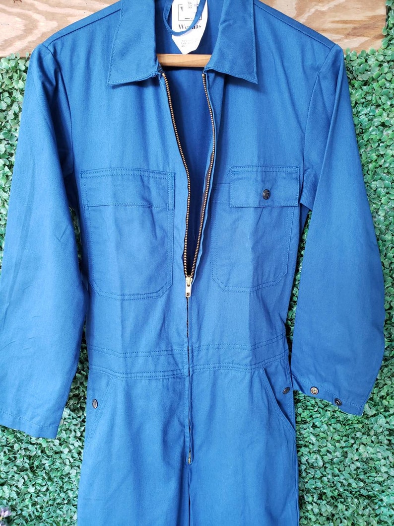 May include: A blue denim jumpsuit with a zipper closure and two pockets on the front. The jumpsuit has long sleeves and a collared neckline.