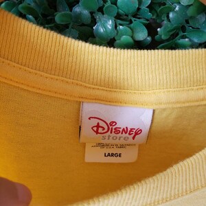 May include: Yellow t-shirt with a white tag that says "Disney Store" and "100% Cotton Assembled in El Salvador of U.S.A. Fabric" and "Large".