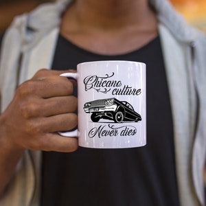 K&ouml;nnte beinhalten: Eine wei&szlig;e Keramiktasse mit einem schwarz-wei&szlig;en Design. Das Design zeigt ein Lowrider-Auto mit dem Text "Chicano culture never dies".