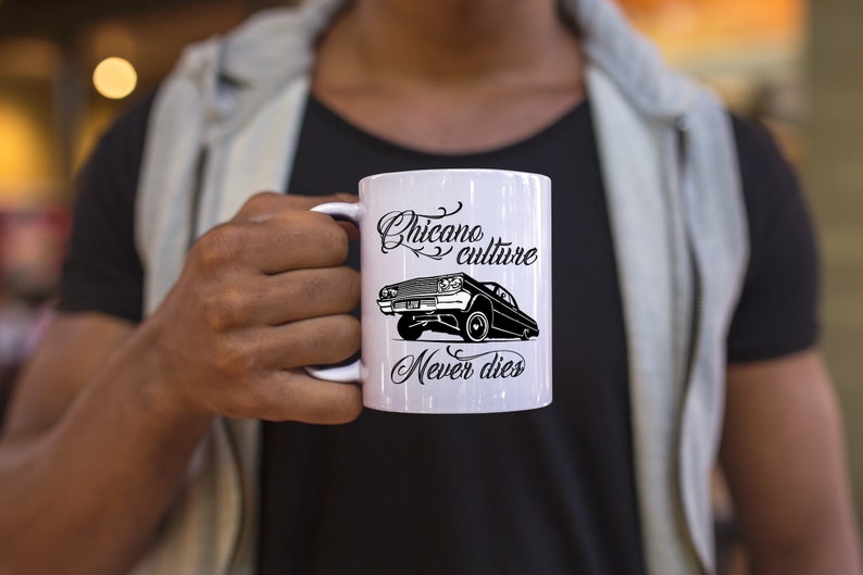 K&ouml;nnte beinhalten: Eine wei&szlig;e Keramiktasse mit einem schwarz-wei&szlig;en Design. Das Design zeigt ein Lowrider-Auto mit dem Text "Chicano culture never dies".