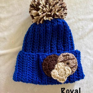May include: A royal blue knit beanie with a large, fluffy pom-pom in shades of brown and beige. The beanie features three crocheted appliques in the shape of cookies. The word "Royal" is printed in black at the bottom.