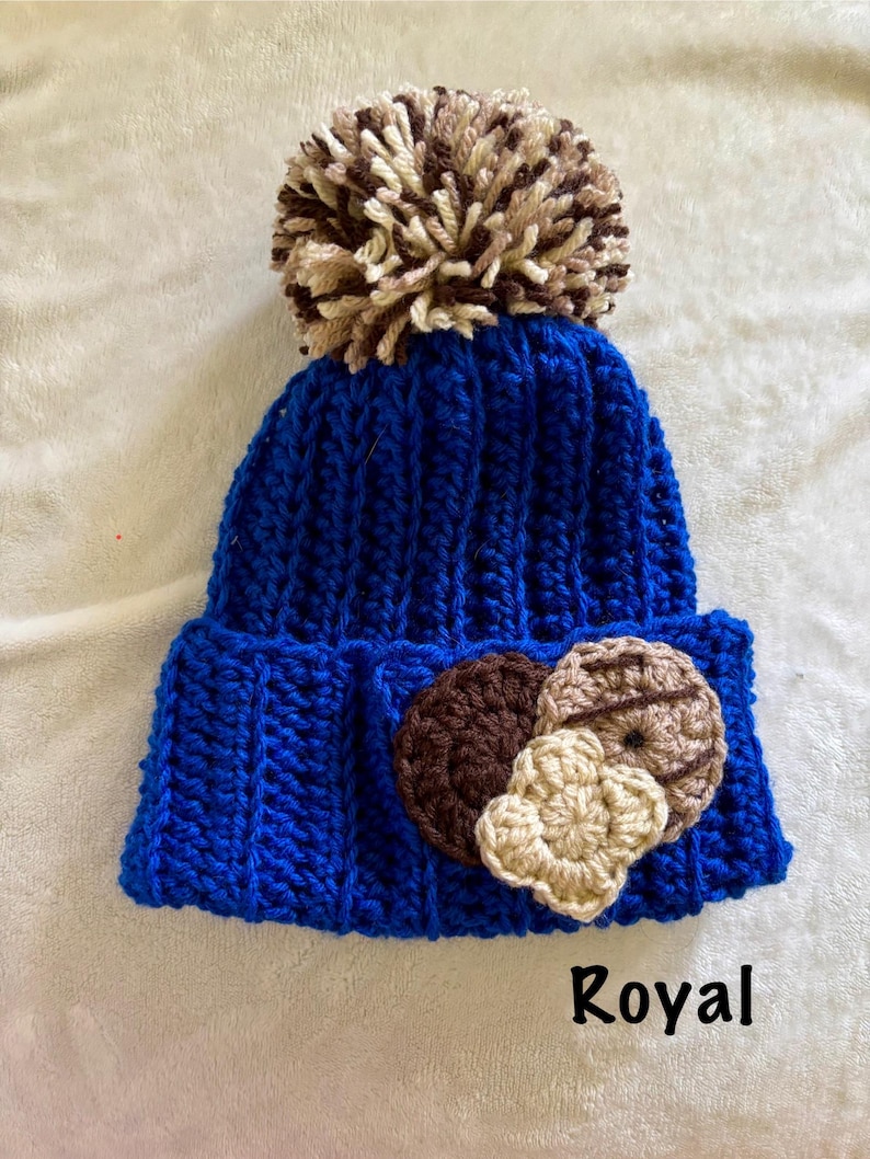 May include: A royal blue knit beanie with a large, fluffy pom-pom in shades of brown and beige. The beanie features three crocheted appliques in the shape of cookies. The word "Royal" is printed in black at the bottom.