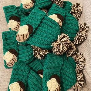 May include: A collection of handmade, green crochet items, possibly cozies or covers, adorned with appliqued cookie designs in cream, brown, and tan. The image also includes several brown and cream pom-poms, suggesting a playful, cozy theme.
