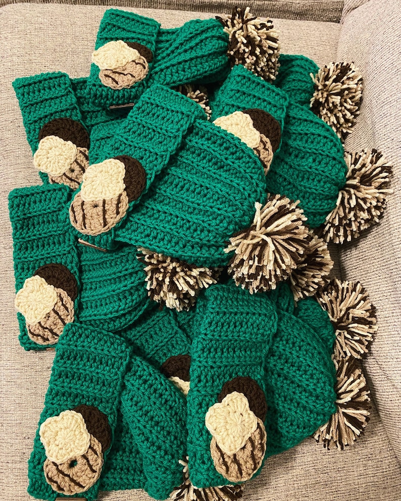 May include: A collection of handmade, green crochet items, possibly cozies or covers, adorned with appliqued cookie designs in cream, brown, and tan. The image also includes several brown and cream pom-poms, suggesting a playful, cozy theme.