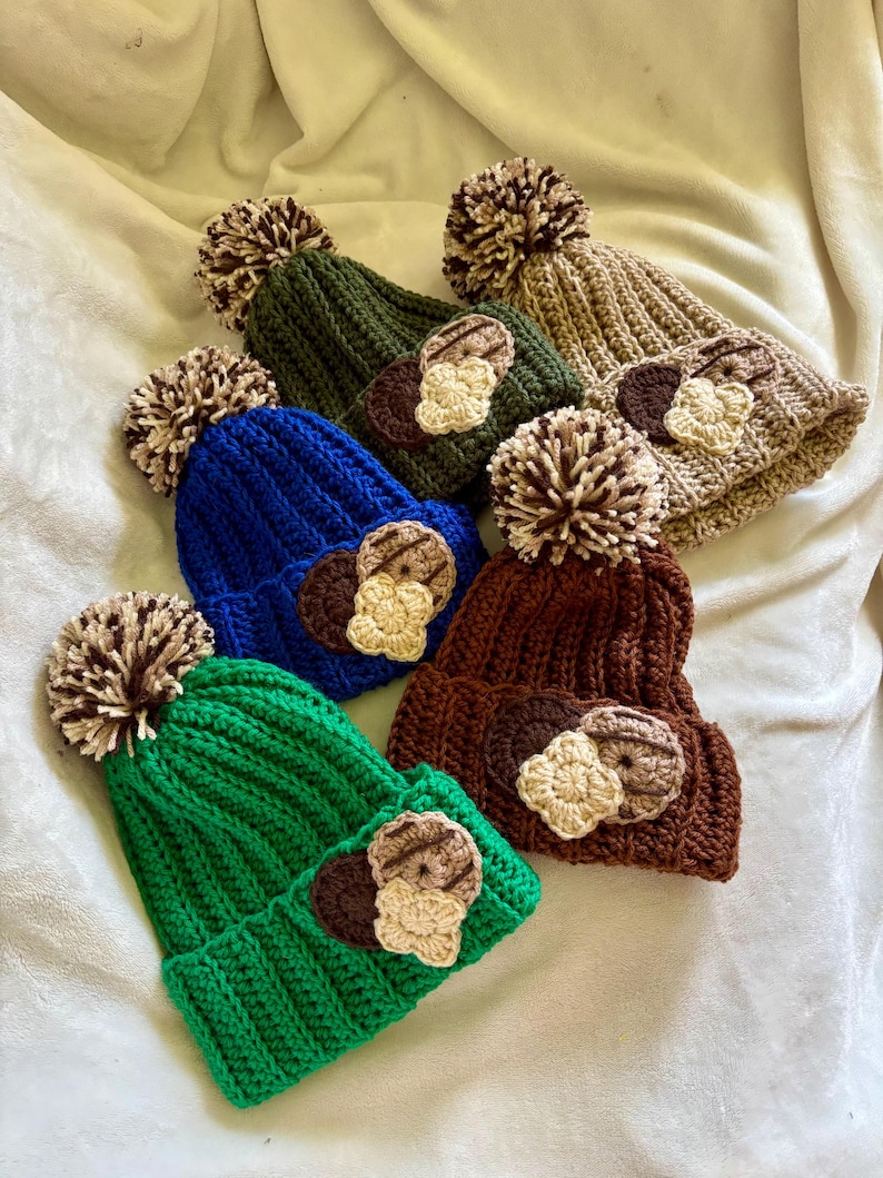 May include: A collection of handmade crochet beanies in various colors, including green, blue, and brown. Each hat features a pom-pom and a decorative applique. The beanies are displayed on a soft, white surface.